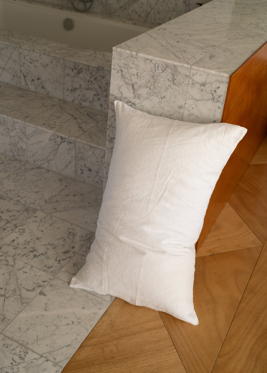 A Pillow slip set - White rests on a crisp pillow against a marble step, beside a wooden panel on a floor with a geometric wood pattern.