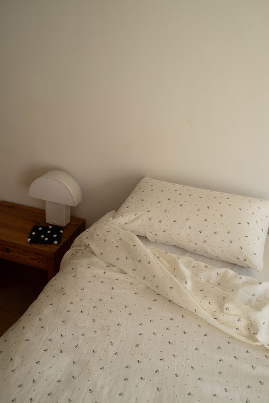 A single bed with a cream sheet and the Duvet cover - vintage floral stands by a wooden nightstand, which holds a white lamp and a black-and-white polka dot coaster, all set against a plain beige wall.