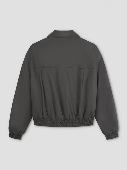 The Shirring Jacket - Slate by Deiji Studios is a dark gray padded jacket with a collar, long sleeves, and an elastic waistband, shown from the back on a plain light background.