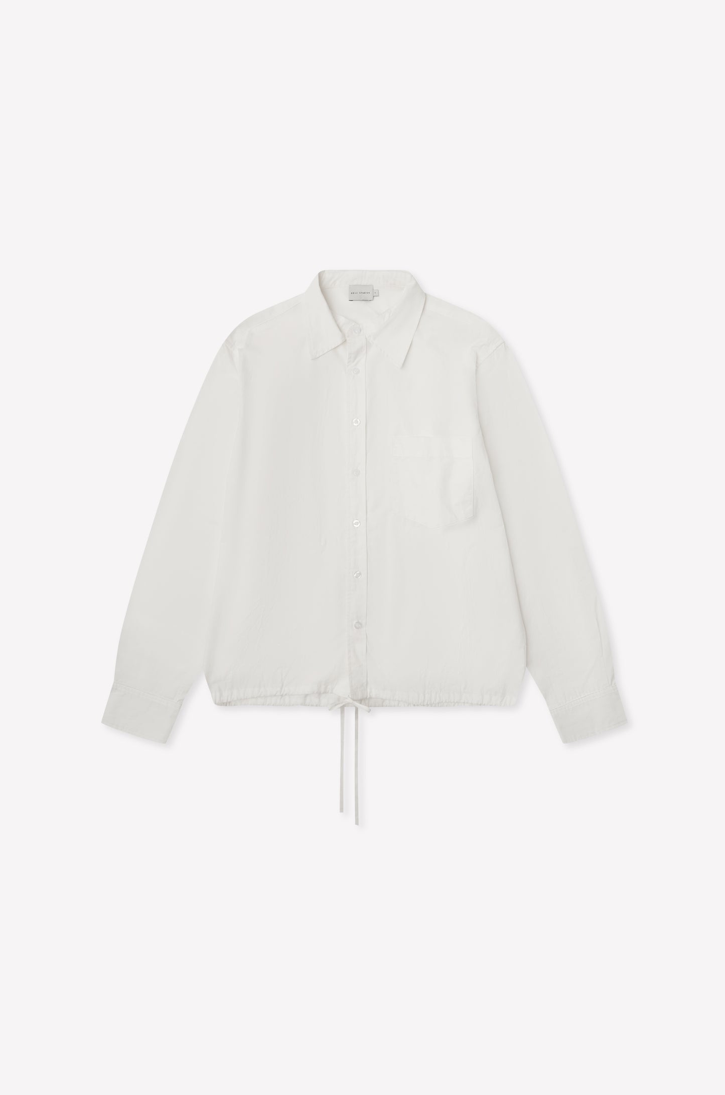 The Stand Shirt - White is a long-sleeved, oversized cotton shirt featuring a single chest pocket, mandarin collar, and drawstring hem, shown on a white background.