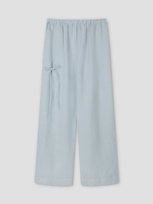 The Eyelet Pant - Soft Blue by Deiji Studios features wide-leg linen, a relaxed ankle length, an elastic waistband, and a small bow tie detail on the left side, displayed against a plain light background.