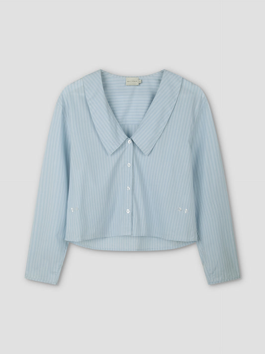 The Waist Button Shirt - Lake Blue Stripe features subtle vertical white stripes, a wide collar, cropped silhouette, long sleeves, and button-up front, displayed on a plain light background.