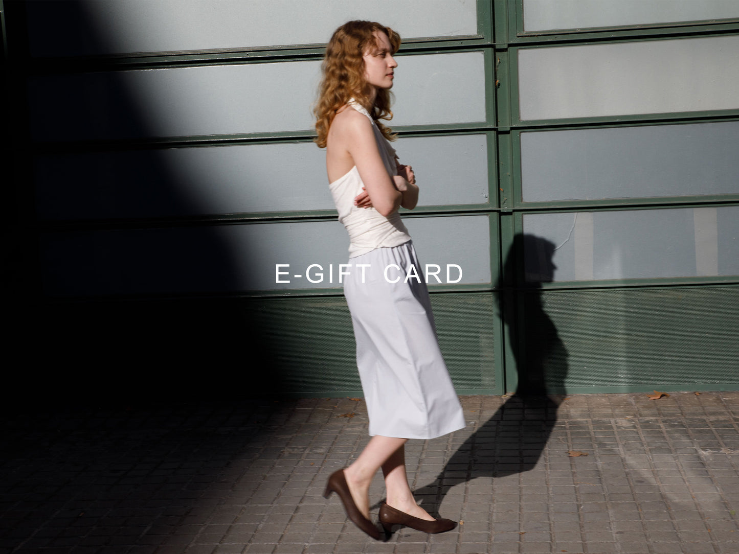 A woman in a white sleeveless top and skirt stands on a sunlit sidewalk by a green glass wall, with "e-gift card—ideal for email delivery of gift cards in Australian Dollars" displayed across the center of the image.