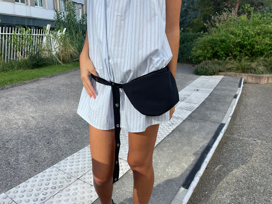 Wearing a light blue, striped Deiji Studios shirt dress and the Snap Belt Bag - Black—an essential everyday accessory—a person stands on a sidewalk near greenery and a white building.