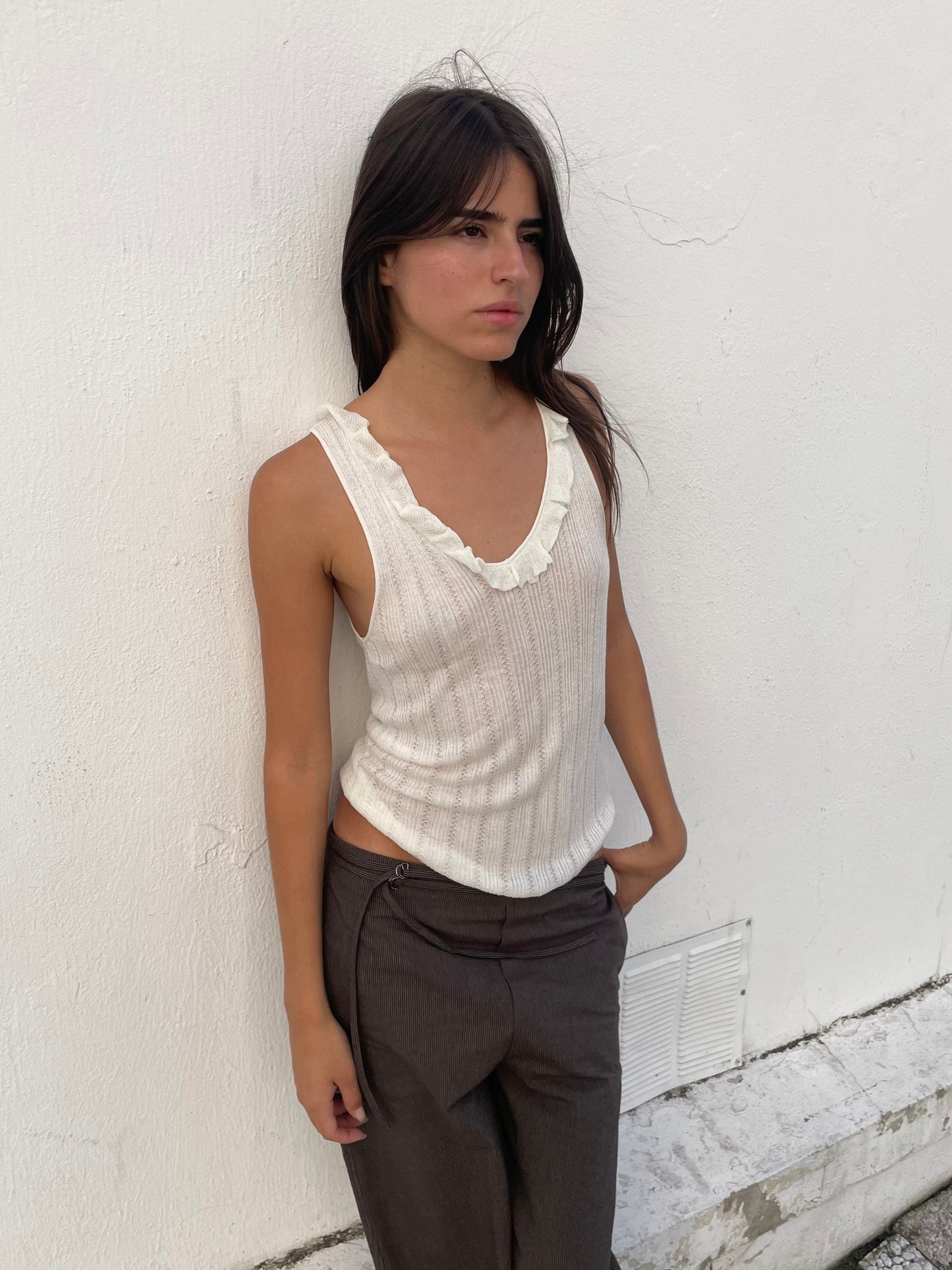 A woman with long dark hair stands against a white wall, wearing the Neck Ruffle Knit - White sleeveless ecoviscose top and dark gray pants. One hand is in her pocket and her expression is neutral.