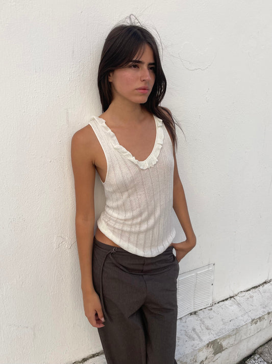 A woman with long dark hair wears the Deiji Studios Neck Ruffle Knit in Off White and gray pants, standing against a white textured wall with her left hand in her pocket, gazing off to the side with a neutral expression.