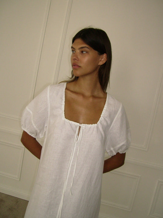 A woman with long dark hair stands before a cream paneled wall, wearing the Curved Seam Midi Dress—a loose-fitting white dress with puffed sleeves and a square neckline—looking to the side with her hands behind her back.