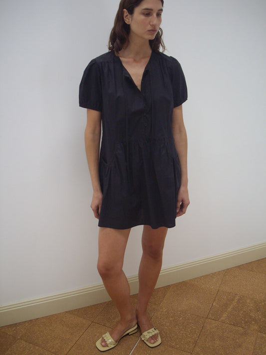A woman with straight brown hair and a neutral expression stands indoors against a plain white wall, wearing Deiji Studios' Pocket Mini Dress in black and light-colored sandals.