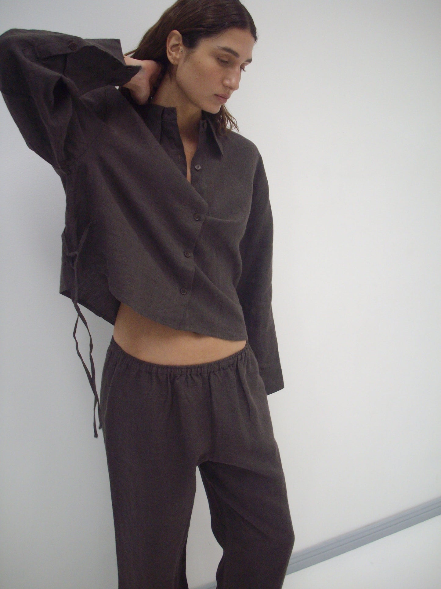 A person wears the Deiji Studios 4 Tie Set - Bark, a loose organic linen outfit in dark brown, standing against a plain white background with one arm raised and head slightly tilted down.