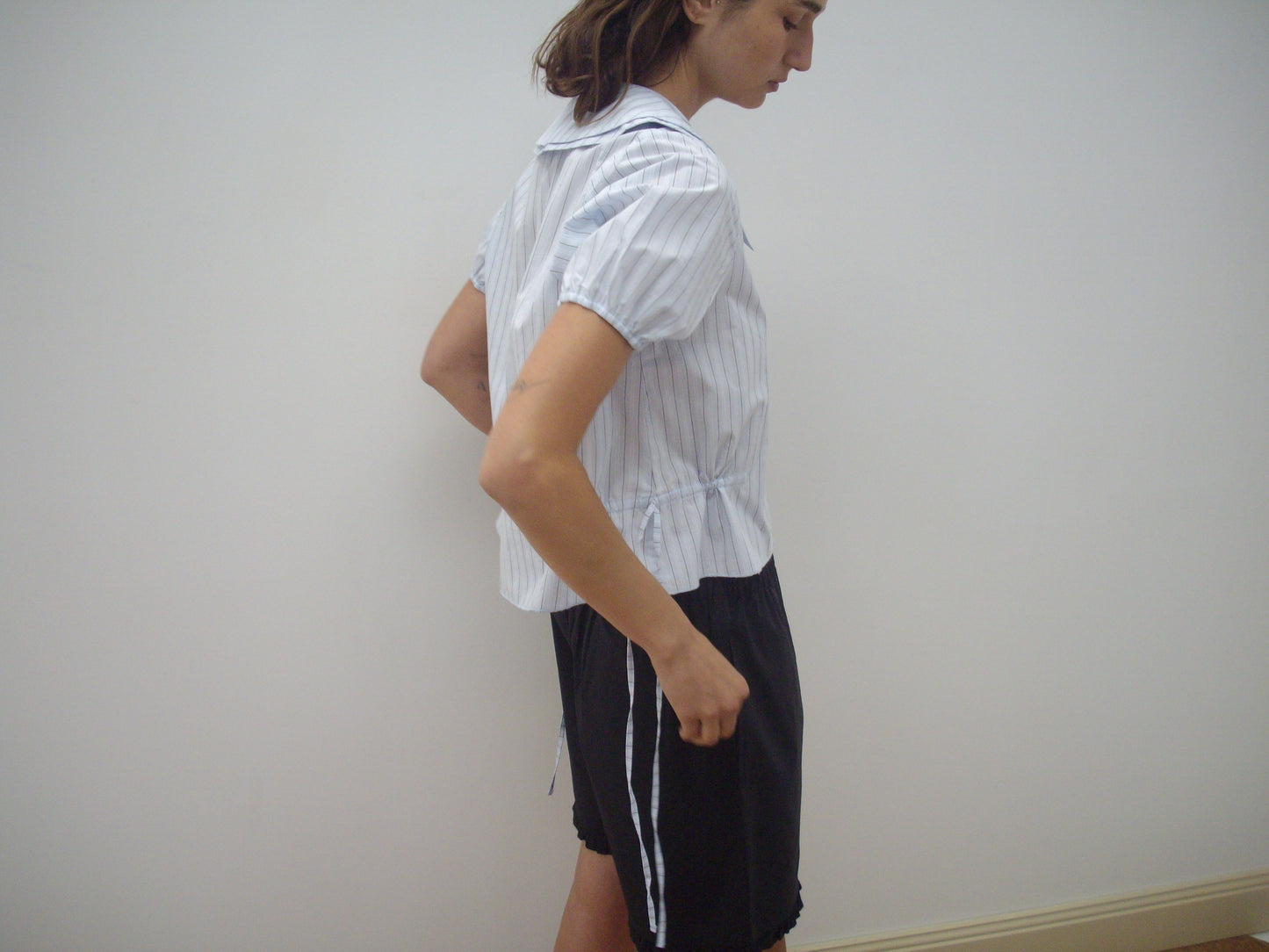 A person stands sideways against a plain white wall, wearing the Deiji Studios Double Collar Shirt - Mineral Stripe and black shorts with white side stripes.