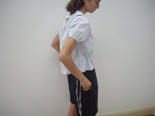 A person stands sideways against a plain white wall, wearing the Double Collar Shirt - Mineral Stripe by Deiji Studios and loose black shorts with side drawstrings.