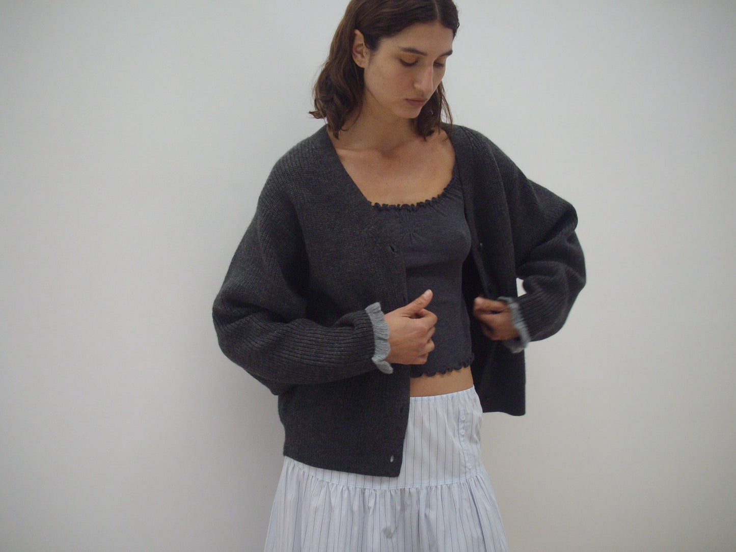 A woman stands against a white background, wearing the Flounce Cardigan - Charcoal over a matching gray top and a light pleated skirt. She looks down, adjusting her cardigan with both hands.