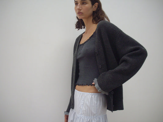 A woman with shoulder-length brown hair and a relaxed expression stands against a white background, wearing the Flounce Cardigan - Charcoal over a gray top and a white skirt with subtle vertical stripes.