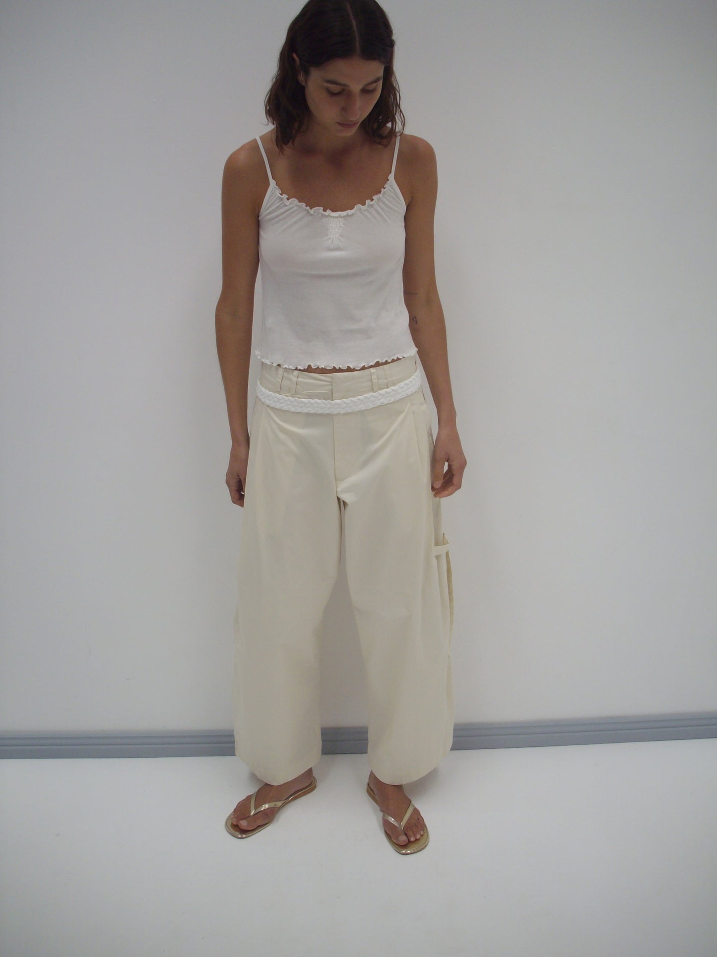 A woman stands indoors against a plain white wall, wearing Deiji Studios’ Tab Pant in off-white, paired with a white camisole and gold sandals. She looks down with her hands by her sides.
