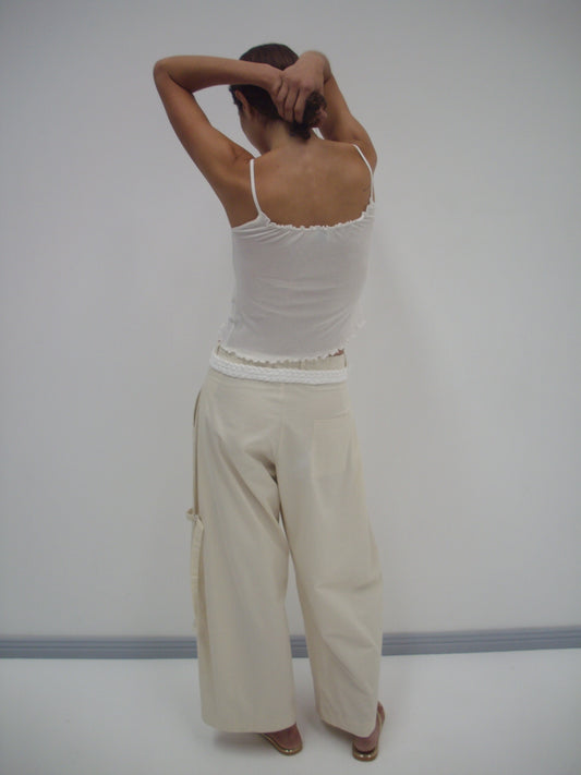 A person stands facing a white wall with arms raised behind their head, wearing a white tank top and the Tab Pant - Off White. The person's back and relaxed posture are visible.