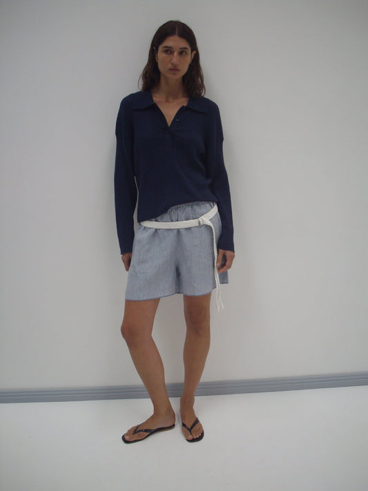 A person with long brown hair and a neutral expression stands against a plain white wall, wearing a Deiji Studios Polo Knit - Dark Blue, light gray shorts with a white belt, and black flip-flops.