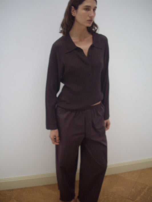 A woman with brown hair stands indoors against a white wall, wearing the Polo Knit - Dark Almond long sleeve top and matching brown pants. She gazes slightly to her left.
