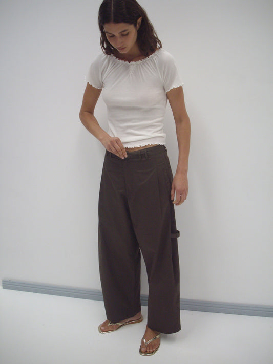 A person wearing a fitted white short-sleeve top, the Tab Pant in dark brown, and sandals stands against a plain white background, looking down and adjusting their pants.