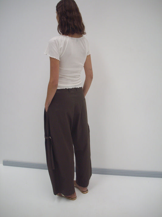 A person with wavy brown hair stands facing away, wearing a white short-sleeve top and the Tab Pant - Dark Brown, organic cotton wide leg trousers with pockets. The background is plain white with a gray baseboard.