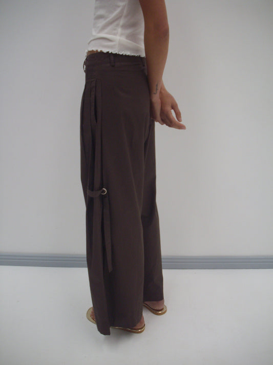 A person stands sideways against a plain white background, wearing the Tab Pant - Dark Brown with a strap detail and a loose fit, paired with a white short-sleeved top.