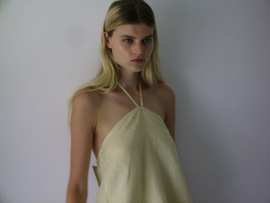 A young woman with long blonde hair wears a Butter Yellow Linen Wrap Back Top by Deiji Studios, standing against a plain white background and looking slightly to the side with a neutral expression.