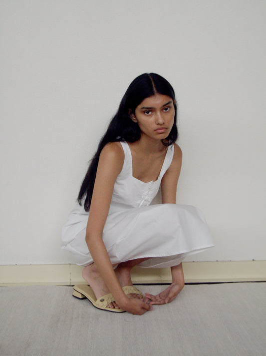 A young woman with long black hair crouches against a white wall, wearing the Deiji Studios Eyelet Dress - White and cream floral slide sandals.