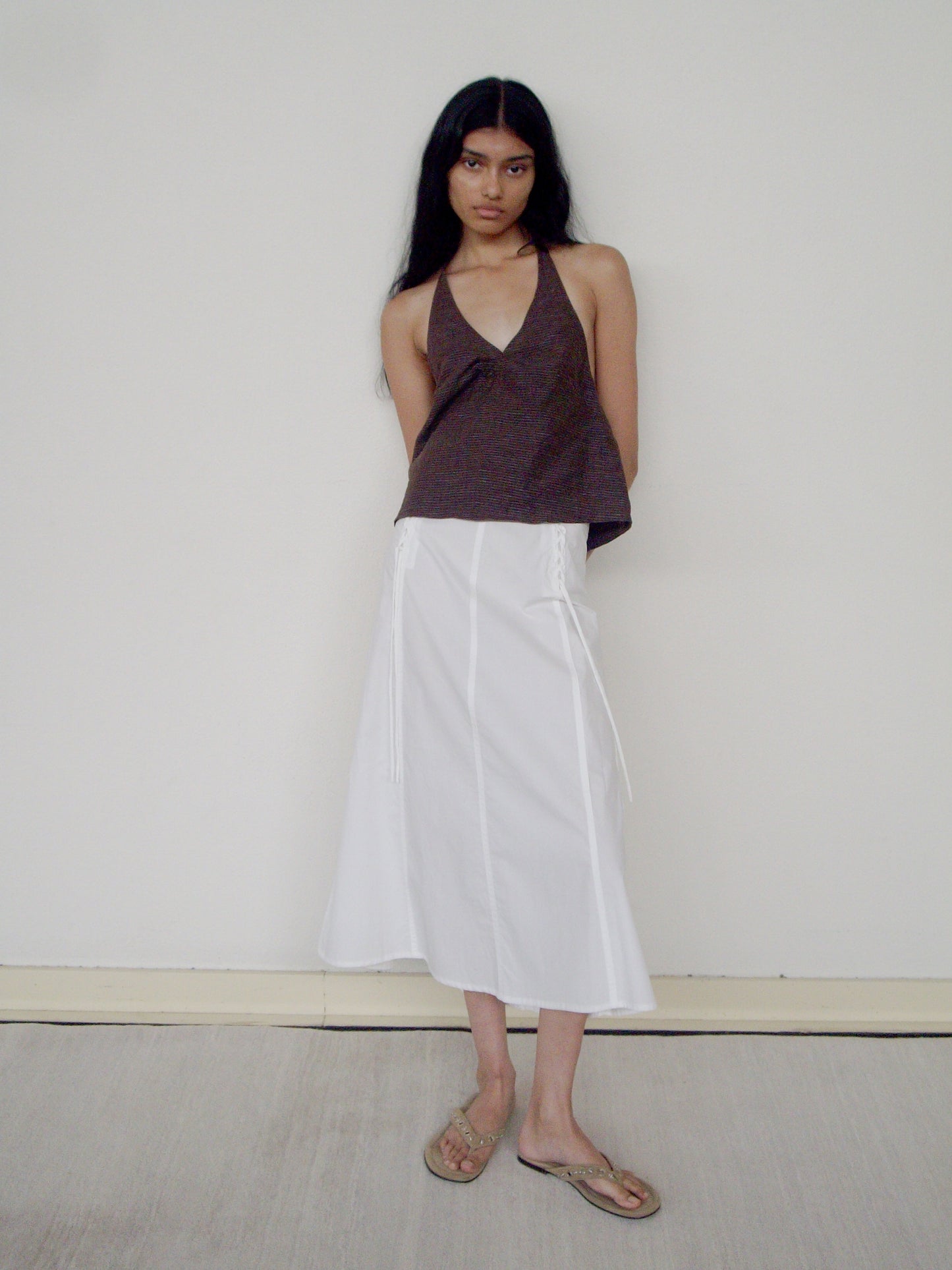 A person with long dark hair stands against a plain wall, wearing a sleeveless brown halter top, the Deiji Studios Double Lace Up skirt in white, and tan sandals. Their arms are behind their back and they have a neutral expression.