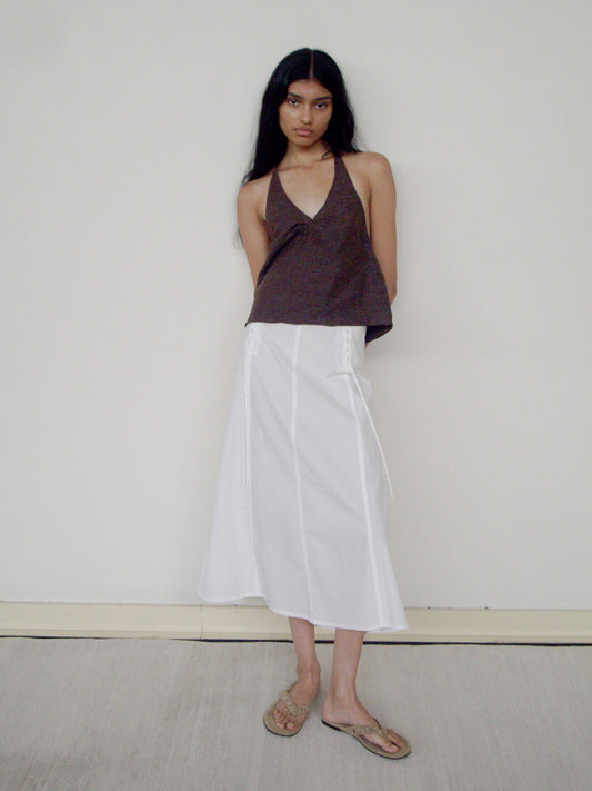 A woman with long dark hair stands against a plain wall, wearing a brown halter top, beige sandals, and the Double Lace Up Skirt—a white midi skirt with side ties. Her arms are behind her back as she looks directly at the camera.