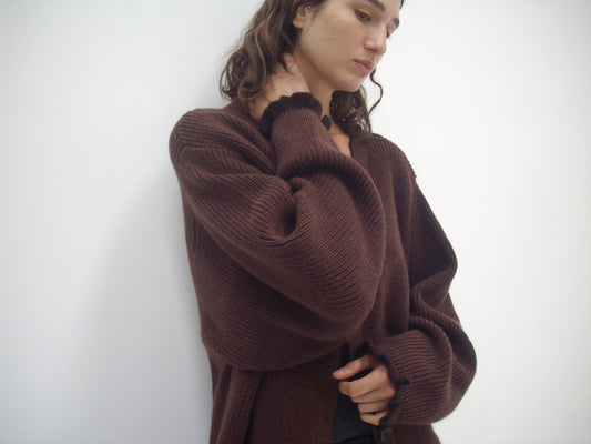 A person with wavy brown hair wears the Flounce Cardigan - Burgundy from Deiji Studios over a black shirt, standing against a plain white background and thoughtfully looking down with one hand touching their neck.