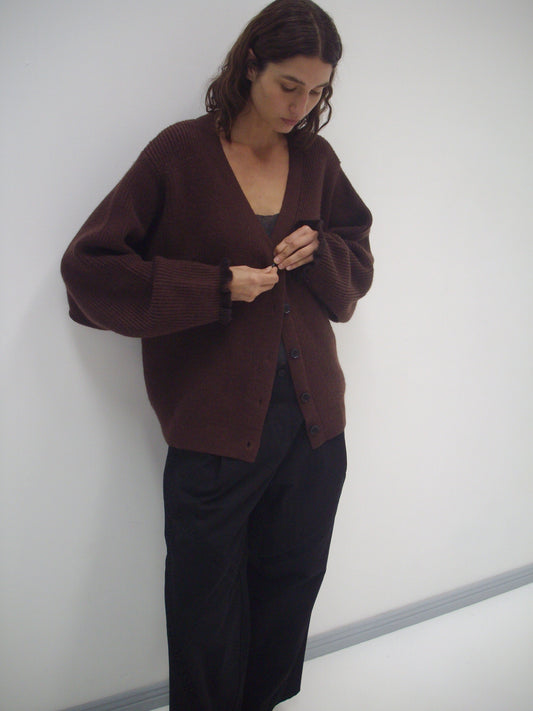 A person with shoulder-length brown hair stands against a white wall, looking down while buttoning the Flounce Cardigan - Burgundy over a dark top and wide-leg black pants.