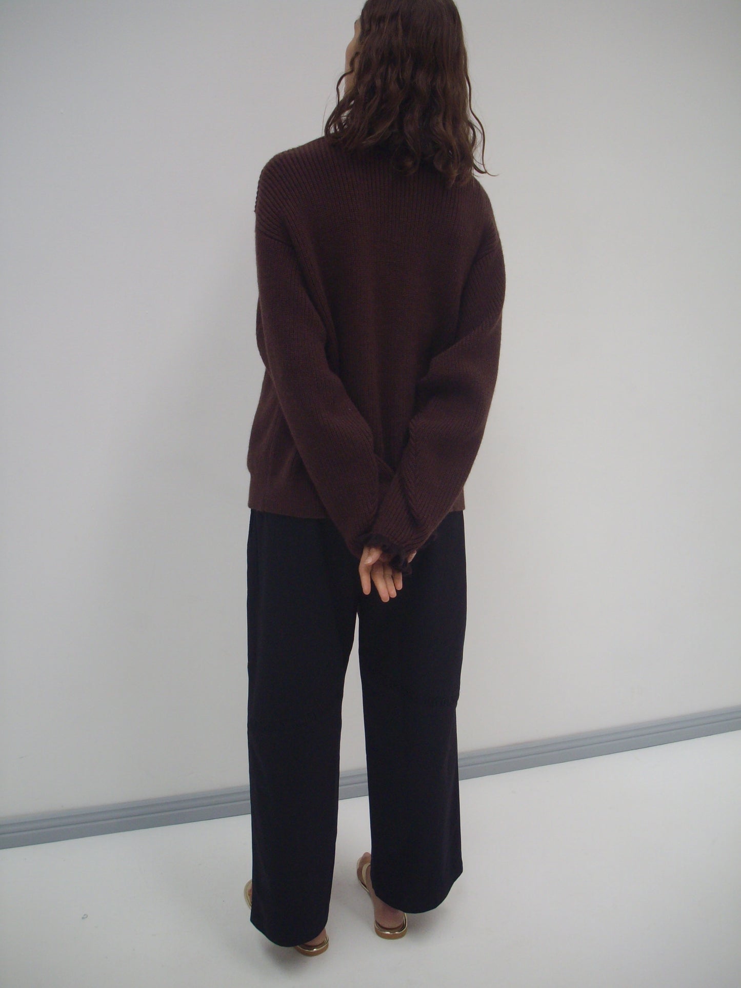 A person with wavy brown hair faces a blank wall, wearing a loose Flounce Cardigan - Burgundy from Deiji Studios, black wide-leg pants, and brown sandals. Their hands are clasped behind their back.