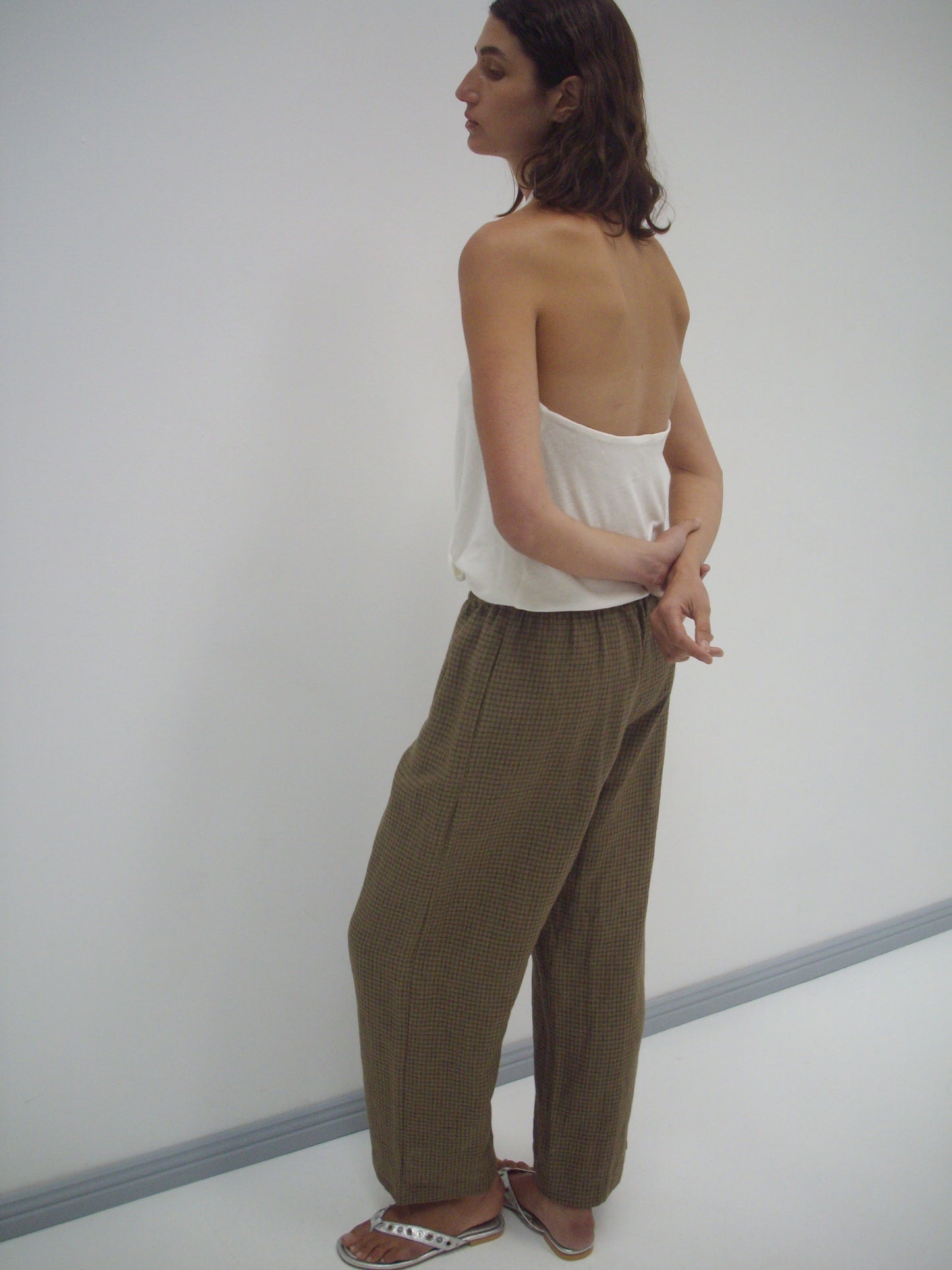 A woman stands sideways in a minimalist room, wearing a strapless white top and the Linen Ease Trouser - Pine Check. Her arms are crossed behind her back as she gazes slightly upward in effortless style.