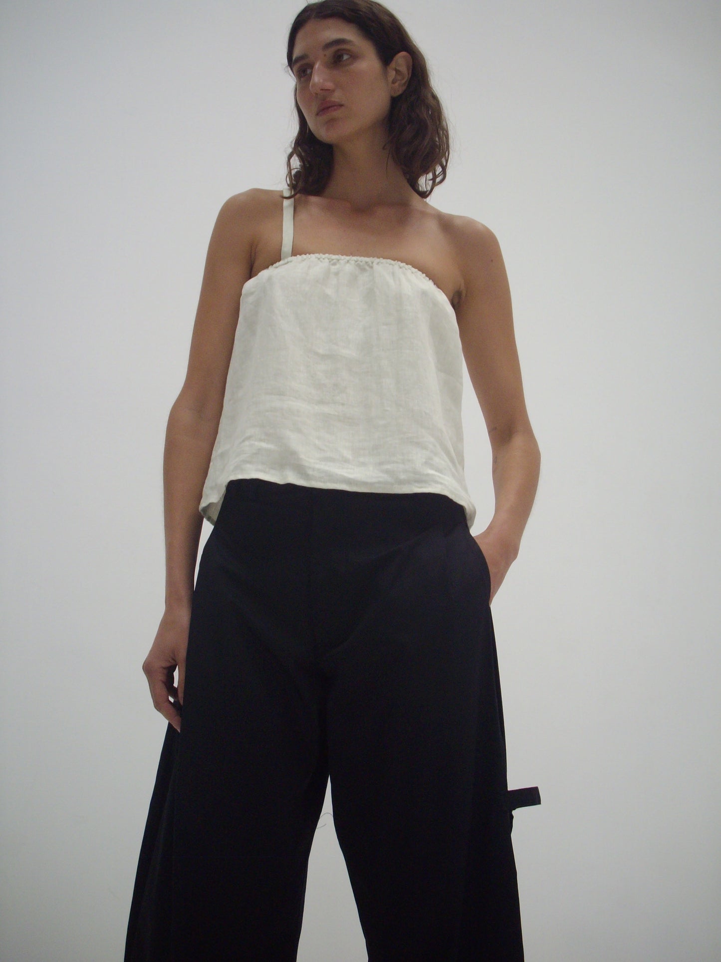 A person with long brown hair wears a white strapless top and Deiji Studios Tab Pant - Black, standing against a plain white background with hands in pockets, looking slightly to the side.