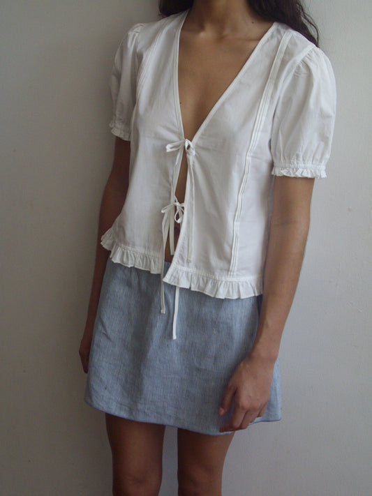 A person stands against a plain wall wearing the Pintuck Top - White by Deiji Studios, featuring front ties and blouson sleeves, paired with a light blue, above-the-knee skirt. Their head is partially out of frame.
