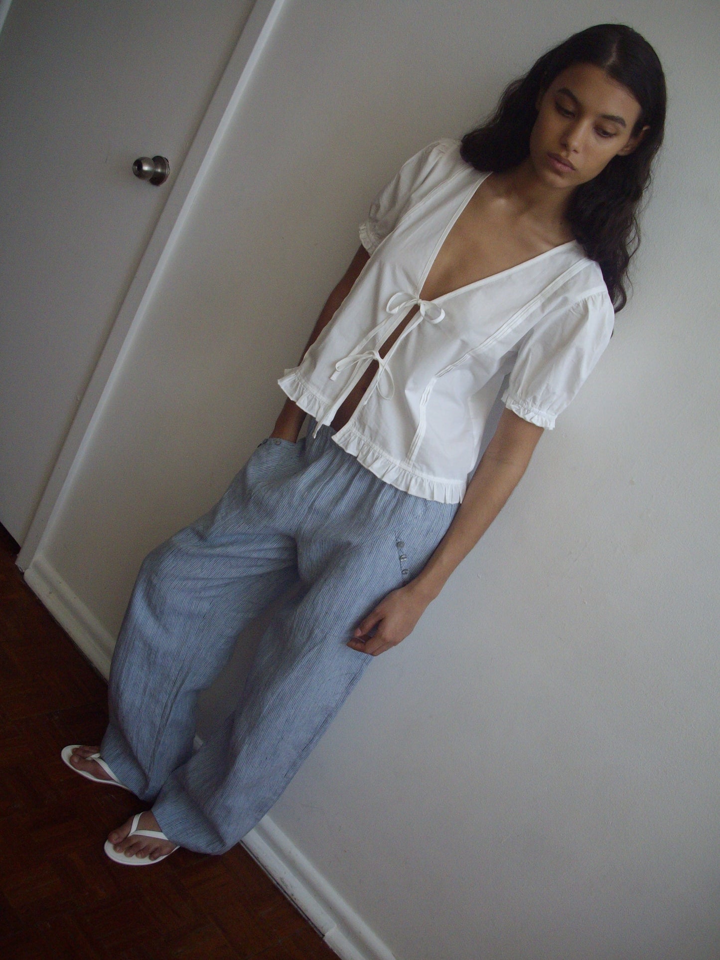 A person with long dark hair stands indoors by a white wall, wearing the Deiji Studios Pintuck Top - White in organic cotton poplin, light blue loose pants, and white sandals. They appear calm and relaxed.
