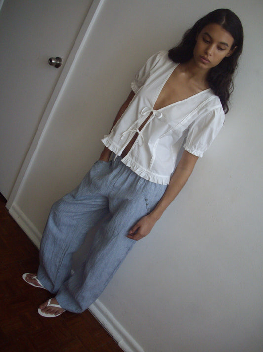 A person with long dark hair stands indoors against a white wall, wearing the Pintuck Top - White by Deiji Studios with blouson sleeves, paired with loose light blue pants and white sandals. They look relaxed, gazing downward.