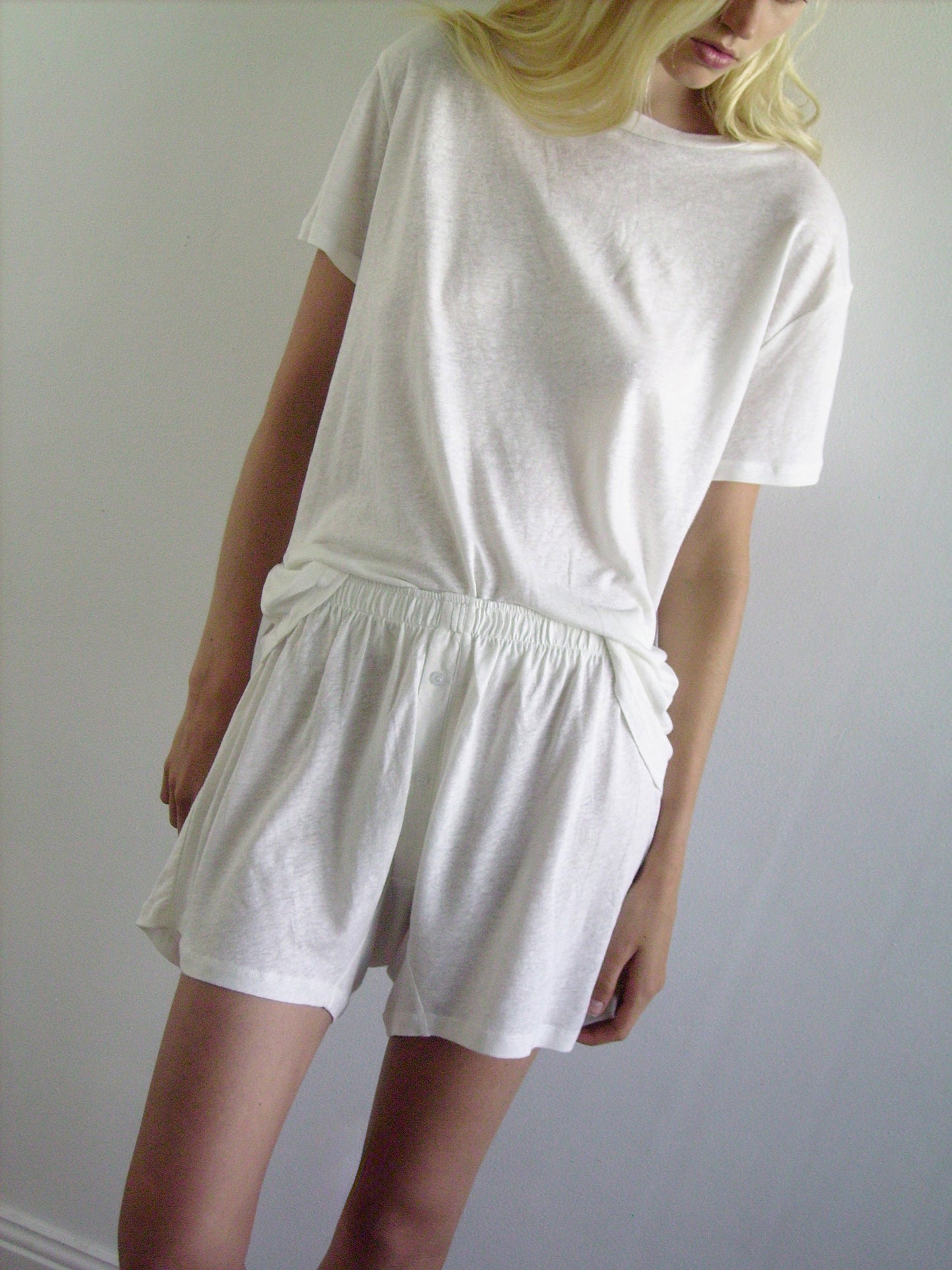A person with long blonde hair wears 0s soft short - ecru and a loose white tee, standing against a plain light wall. Their head is slightly tilted down and to the side, perfectly capturing effortless loungewear style.
