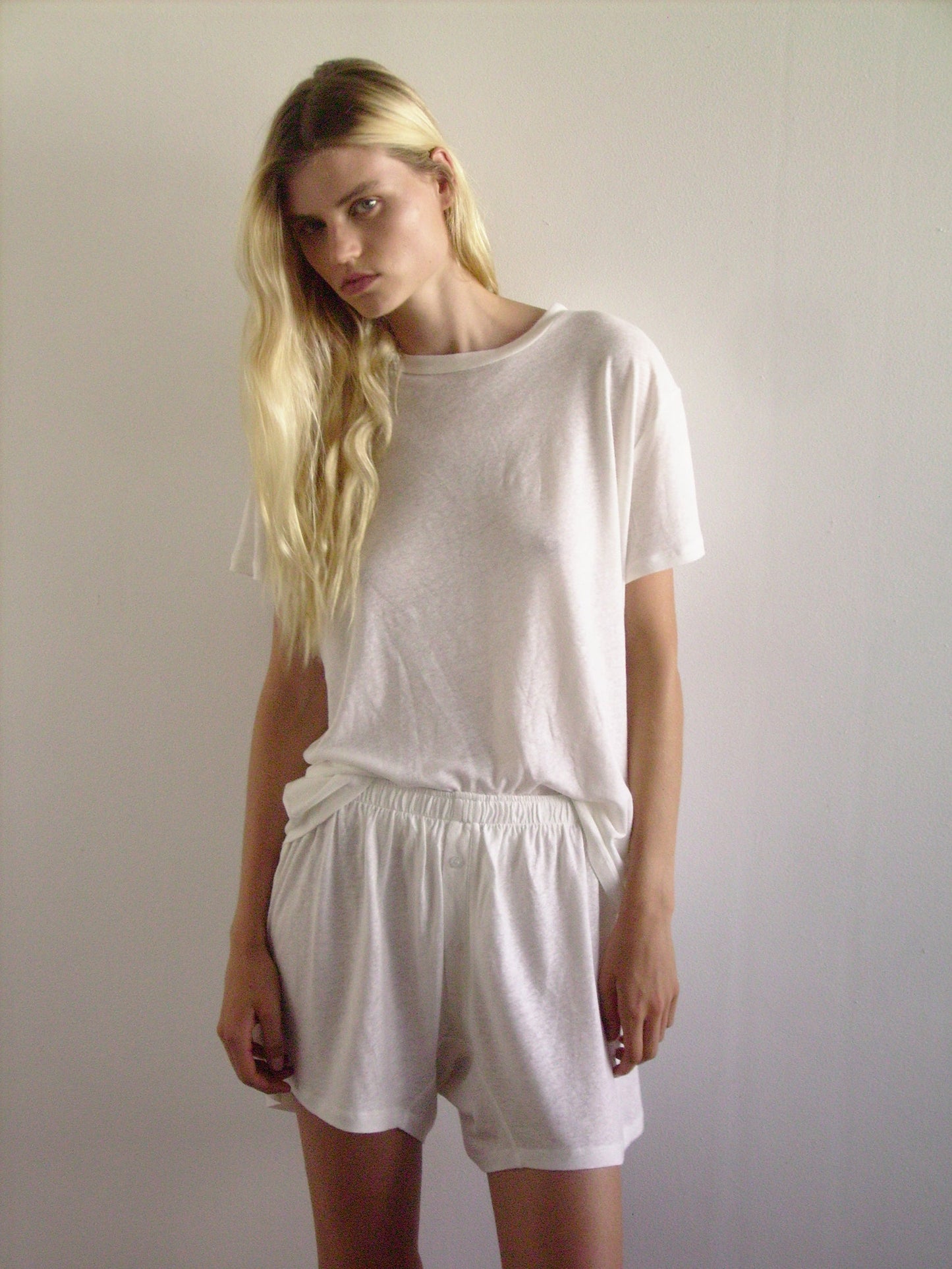 A woman with long blonde hair stands against a plain light wall, wearing 0s soft short - ecru and a loose white tee, looking straight at the camera with a neutral expression.