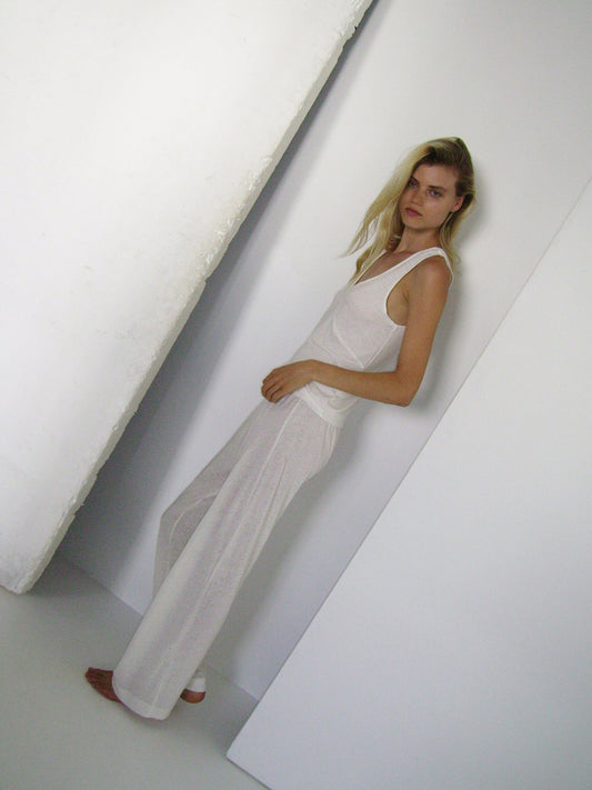 A barefoot woman with long blonde hair stands indoors against a white wall, wearing a loose-fitting Soft Tank and wide-legged white pants in a minimalist, softly lit setting.
