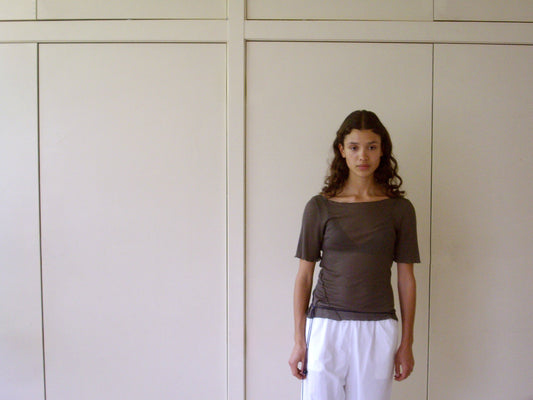 A young woman with long curly hair stands indoors by a beige paneled wall, wearing a Square Knit Tee - Stone from Deiji Studios and loose white pants, looking at the camera with a neutral expression.