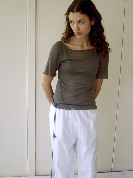 A person with long, curly brown hair wears the Square Knit Tee - Stone by Deiji Studios and loose white pants, standing indoors against a beige wall with hands behind their back, gazing slightly to the side.