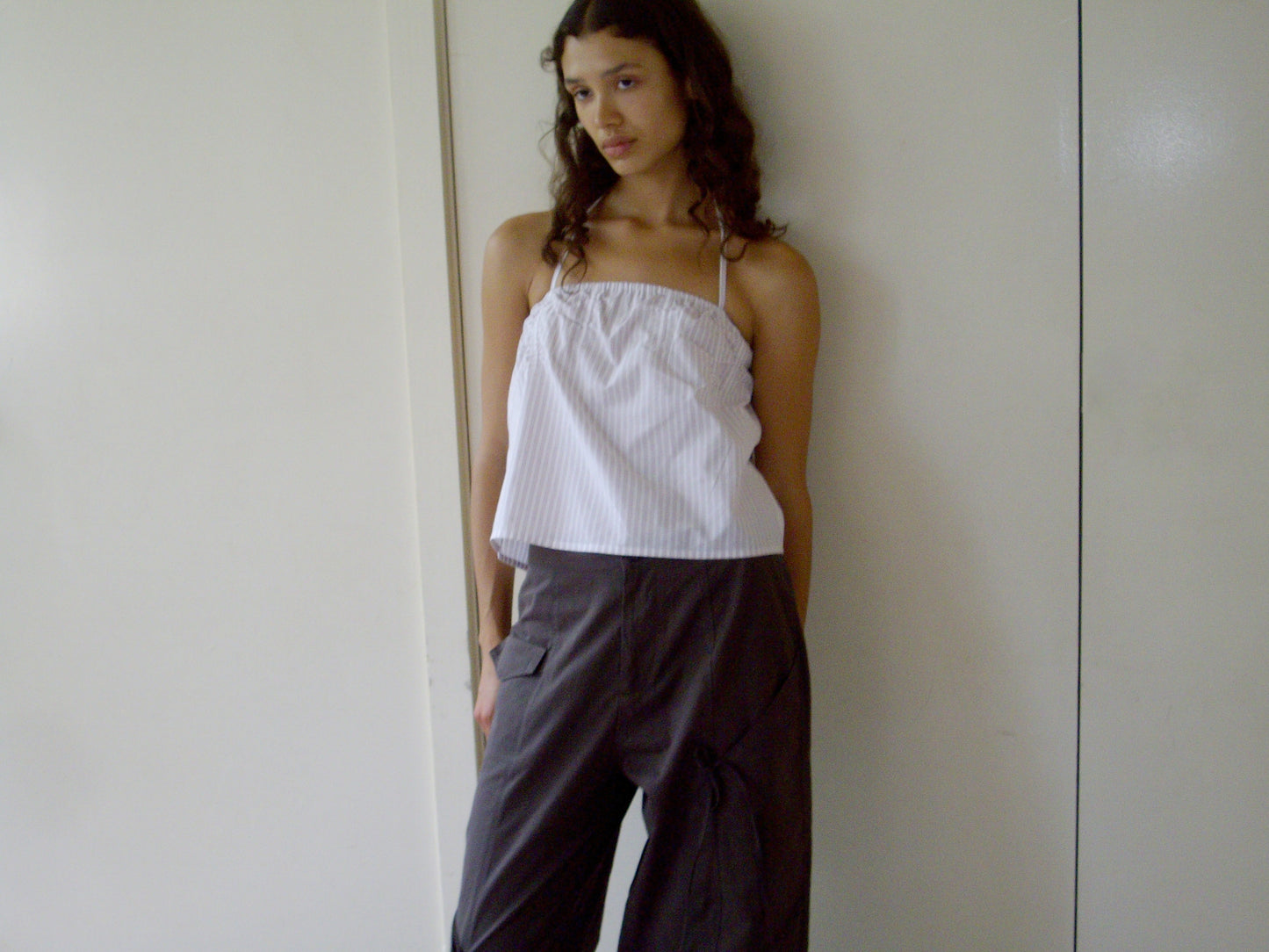 A woman with long curly hair wears the 0 Tube Top - Musk Stripe in a cropped fit with loose dark pants, standing indoors against a plain white wall and looking to the side with a relaxed expression.