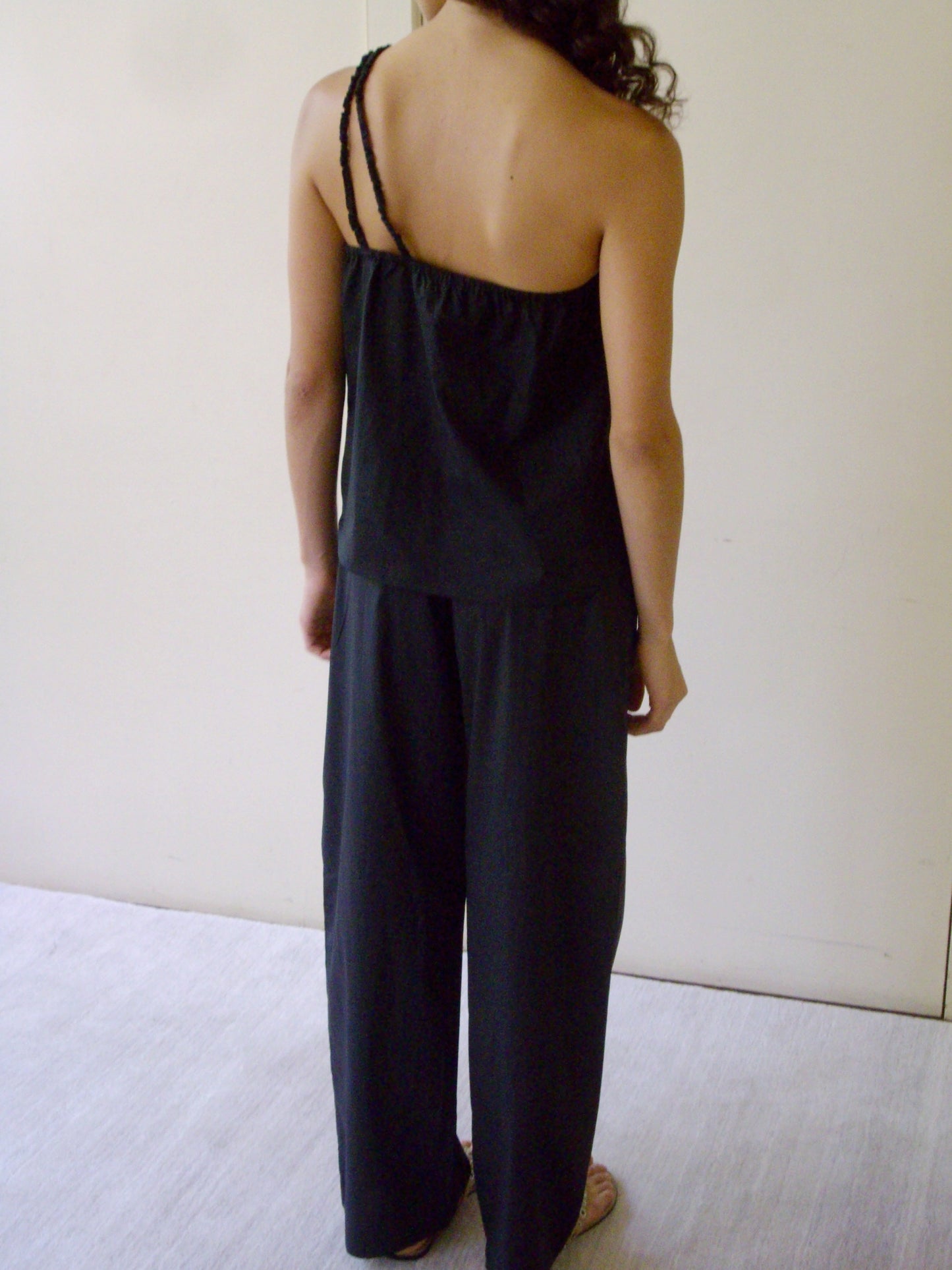 A person with long, wavy hair stands indoors facing away, wearing the Deiji Studios Shoulder Top - Black with uneven straps and loose black pants. The background is plain with light-colored walls and flooring.