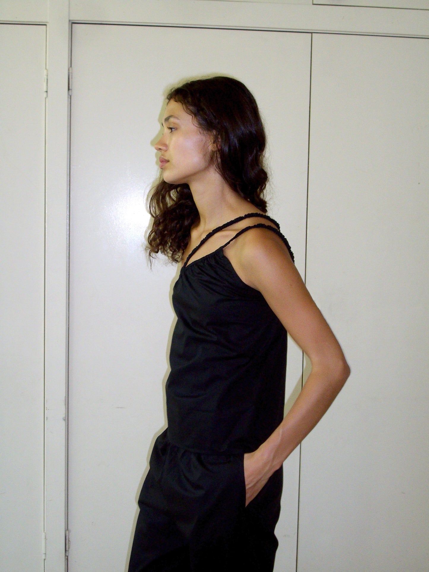 A person with long, wavy brown hair stands in profile against a light wall, wearing Deiji Studios Shoulder Top - Black in organic cotton and matching pants, with one hand in their pocket.