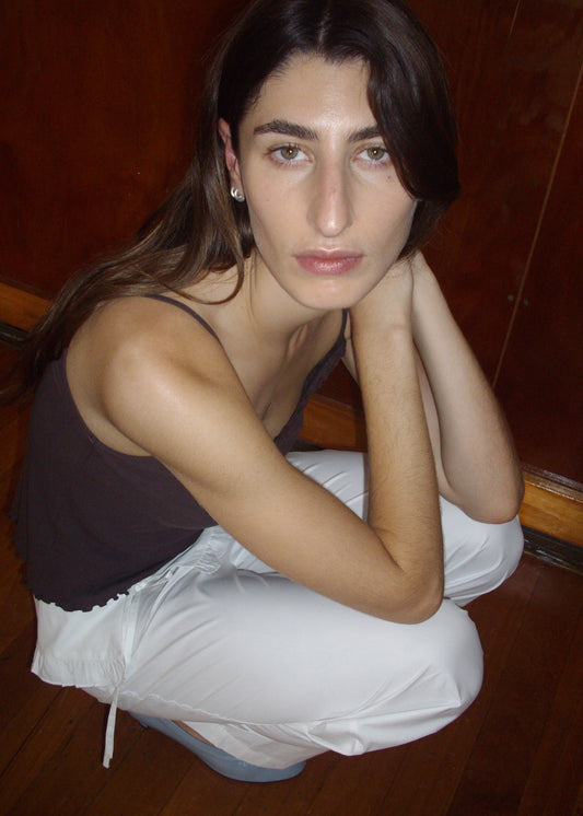 A person with long brown hair and light skin is squatting on a wooden floor, wearing the Cotton Flounce Tank - Brown and white pants, looking up at the camera with a neutral expression.