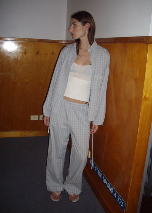 A woman stands indoors in the Side Tie Pant - Blue Check over a white top and sandals, looking to the side near wooden paneled walls and a blue sign on the floor.