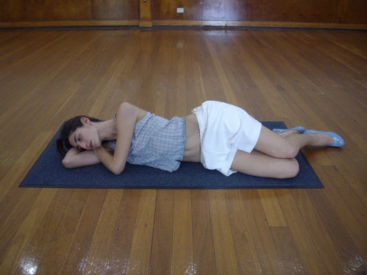 A person lies on their side on a yoga mat in a spacious room, wearing Deiji Studios’ Relax Short - Off White, a sleeveless top, and gray flat shoes.