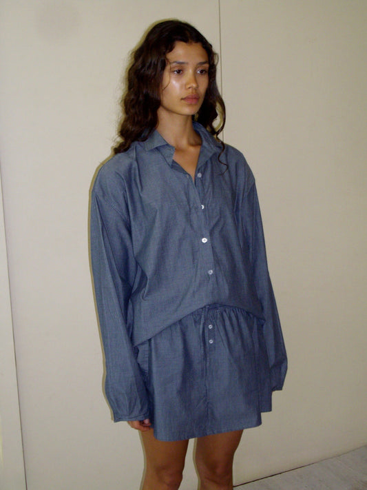 A woman with long, wavy hair stands against a plain wall, wearing the oversized blue day shirt styled as a dress. She has a neutral expression and her hands are by her sides.