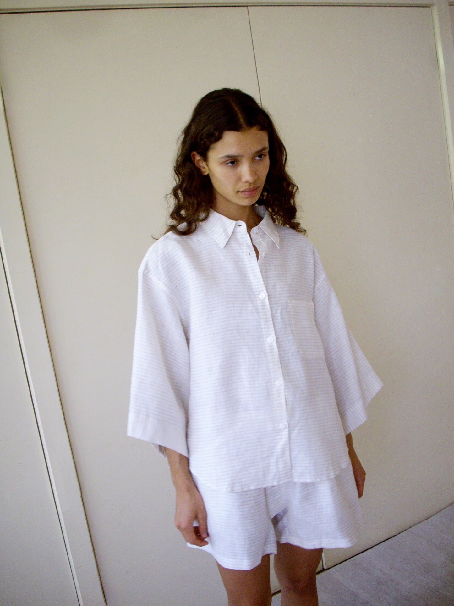 A person with long, wavy hair stands indoors wearing the 03 Set - Linen Story Stripe: a loose-fitting organic linen shirt with wide sleeves and matching boxer-style shorts. Light-colored sliding doors are in the background.