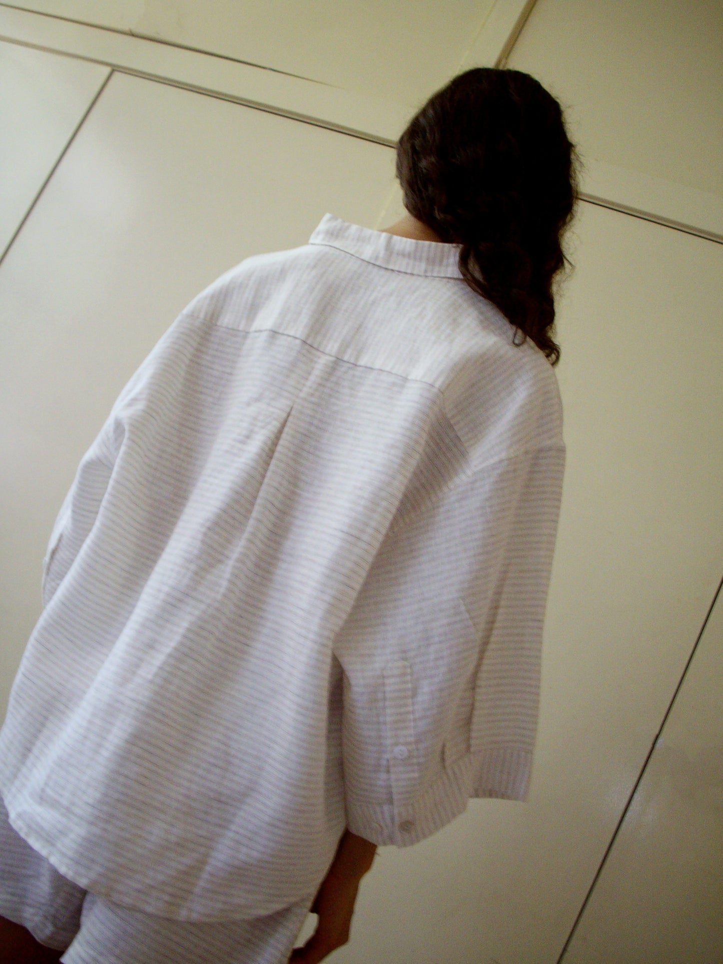 A person with long, dark hair stands facing away, wearing the 03 Set - Linen Story Stripe: a loose, light-striped organic linen shirt and matching boxer-style shorts, in front of a plain light wall or wardrobe.